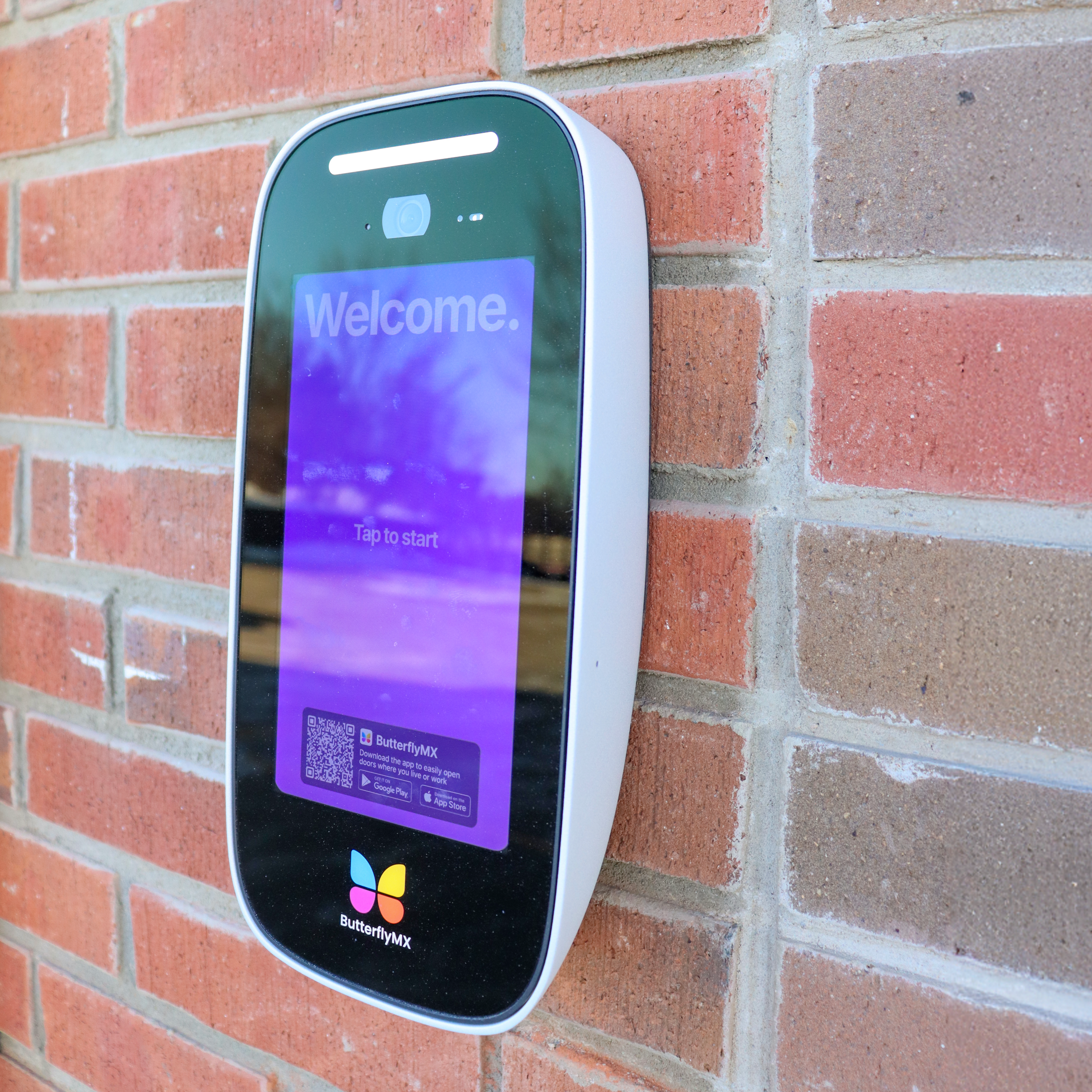 A close-up of a ButterflyMX entry system installed on a brick wall, displaying the "Welcome" message on the screen.