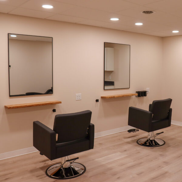 a Double Studio at Riviera Salon Studios equipped with two modern styling stations that include. styling chair, a mirror, a metal styling tool holder, and a wood shelf.