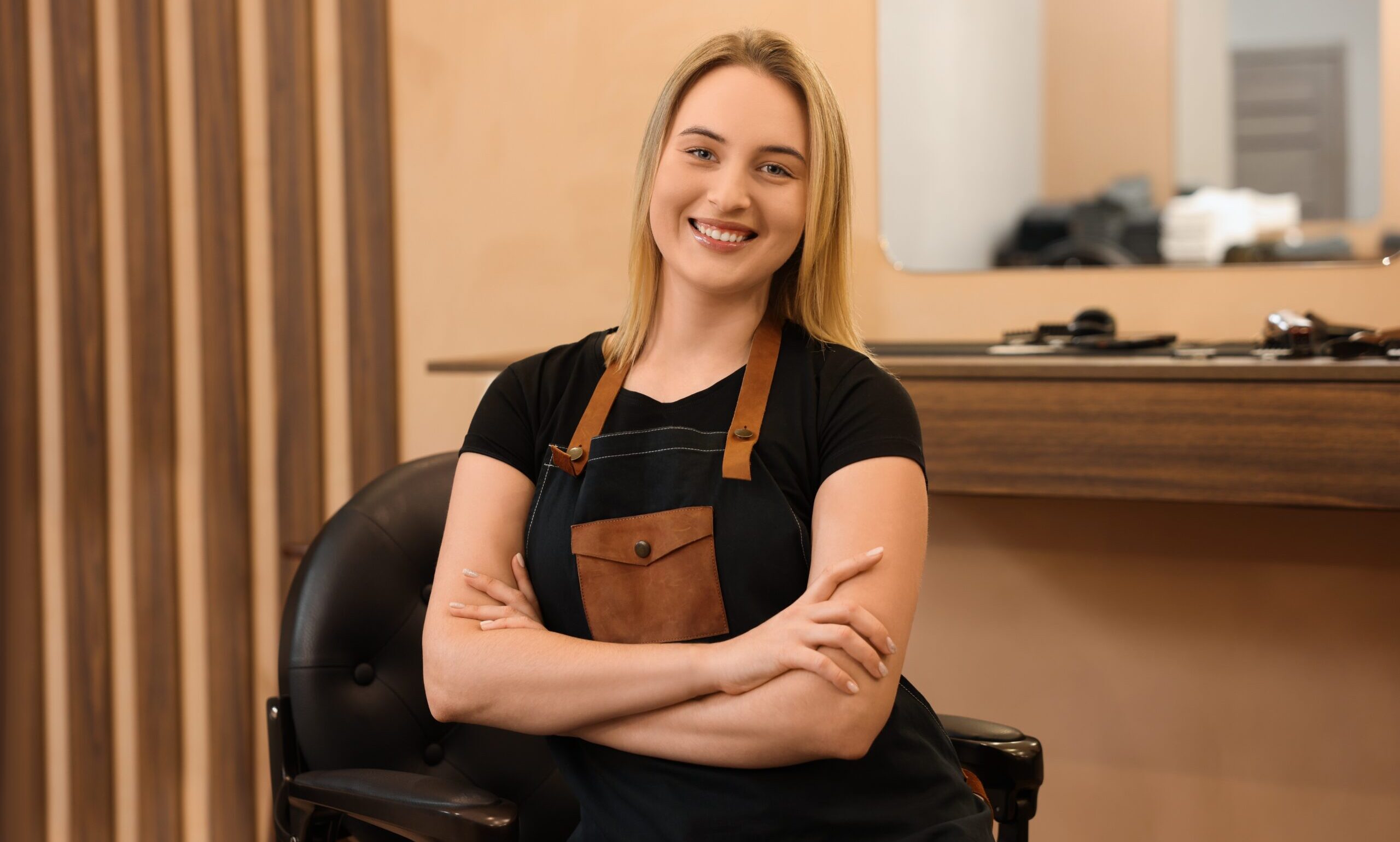Confident young female hairstylist smiling while standing in a salon with arms crossed.