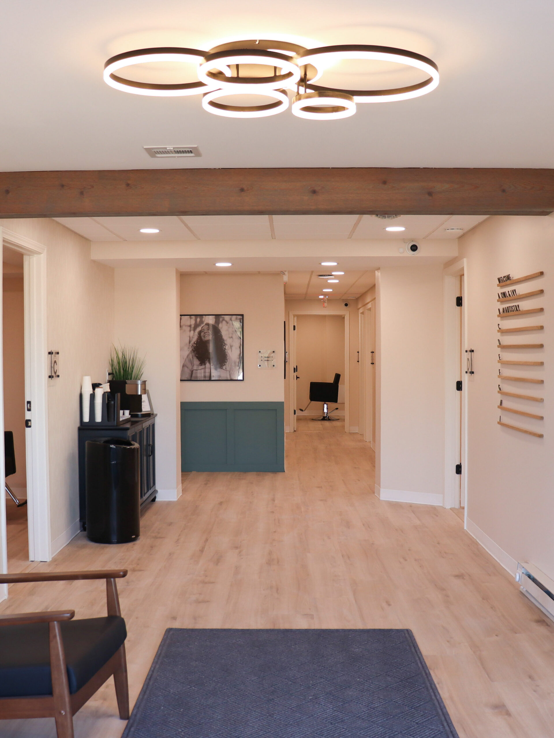 View of the front waiting area from the front door of Riviera Salon Studios, featuring a modern seating area, a stylish chandelier, and a welcoming hallway with studio doors.